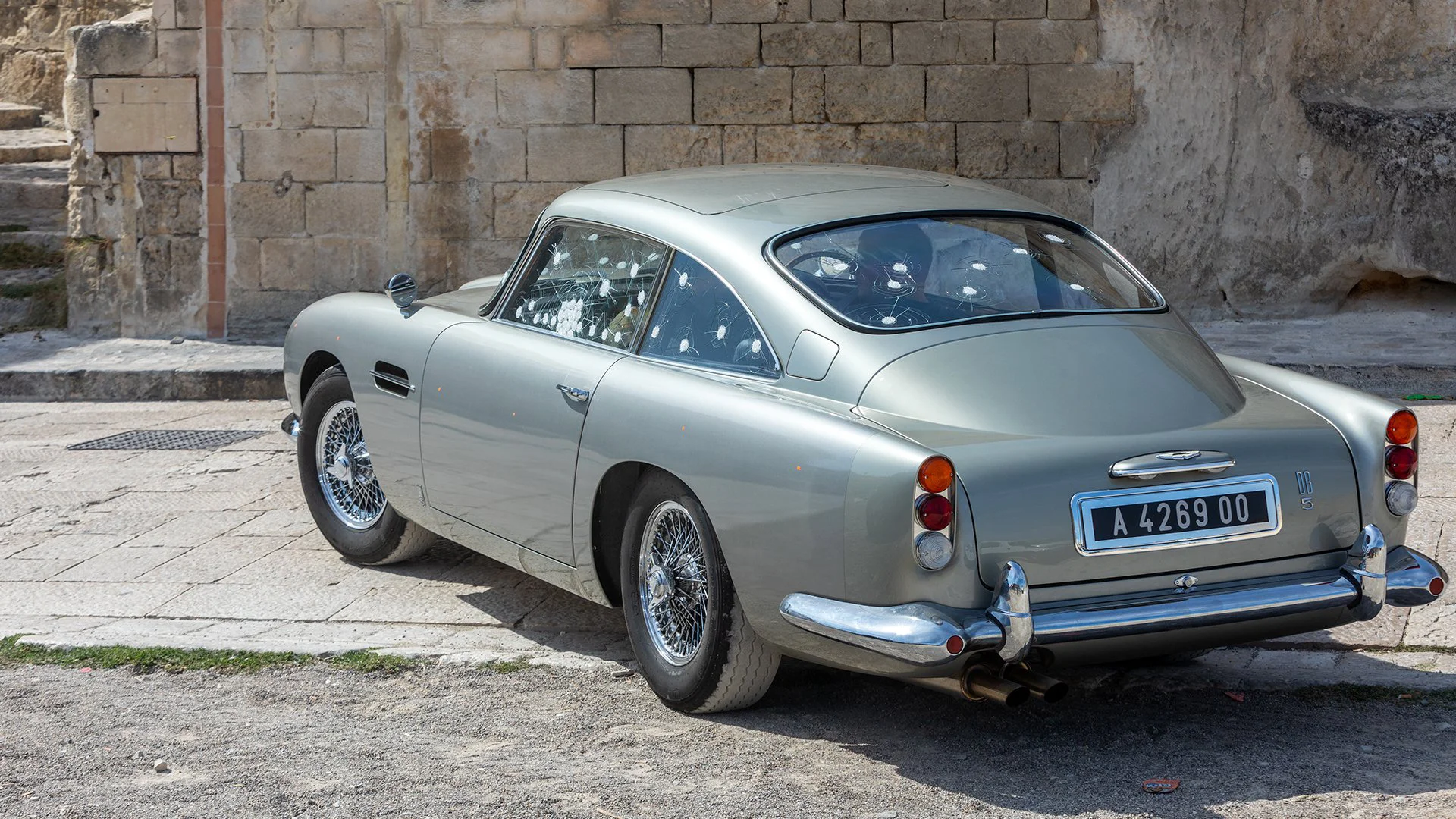 Carro Aston Martin DB5 com marcas de tiros estacionado sobre piso de pedra, como visto na franquia James Bond - exemplo clássico de product placement.