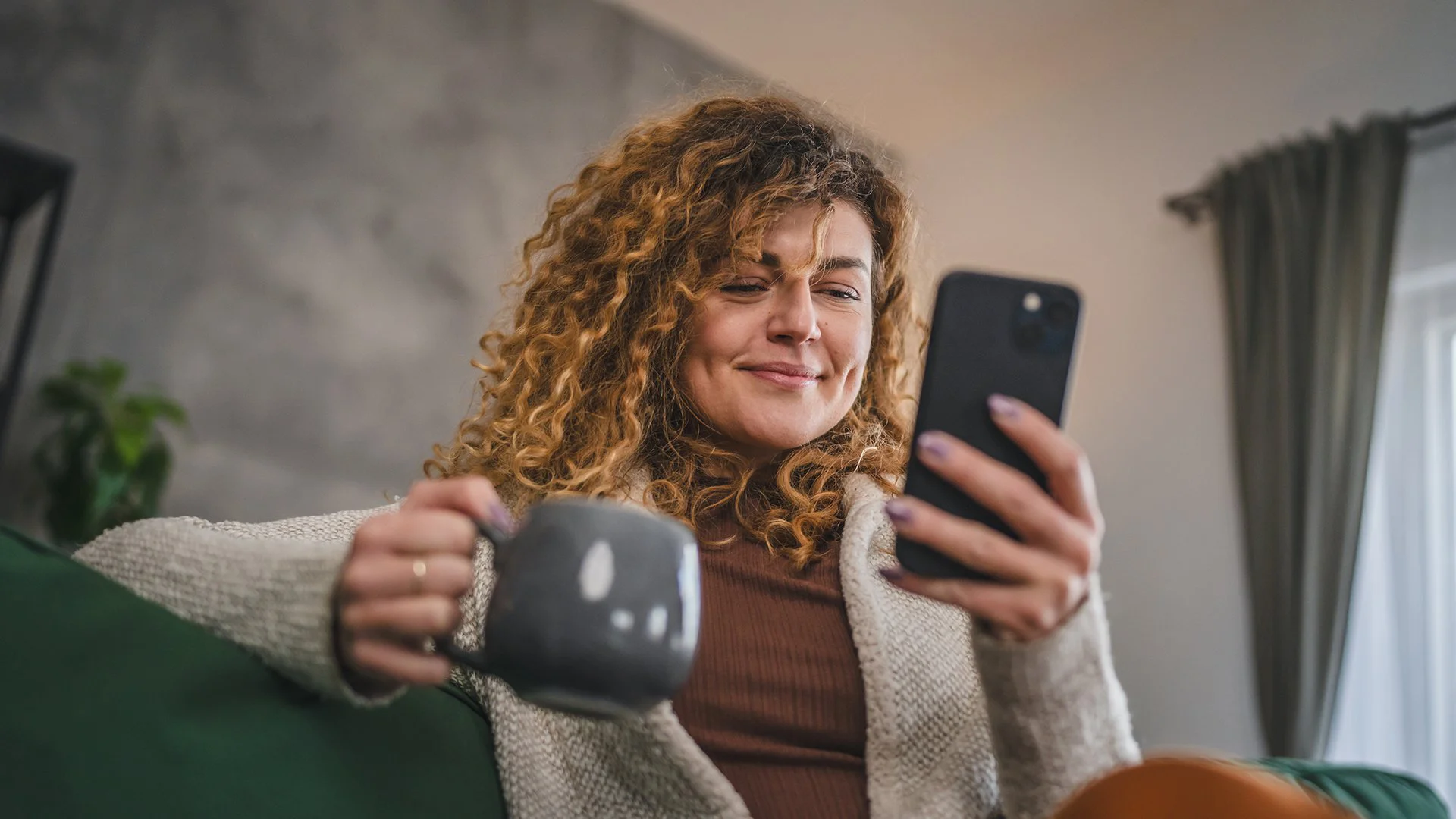 Mulher em ambiente doméstico segurando smartphone e caneca, consumindo conteúdo mobile-first em momento de relaxamento.