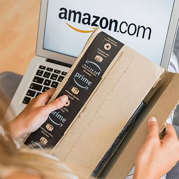 Pessoa abre pacote da Amazon Prime em frente a notebook com o site dessa empresa aberto, representando experiência de consumo digital e branding.