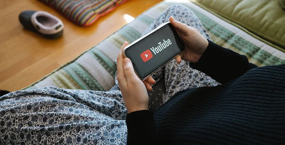 Pessoa segura smartphone com logo do YouTube na tela, sentada em ambiente descontraído, ilustrando consumo de vídeo mobile no marketing digital.