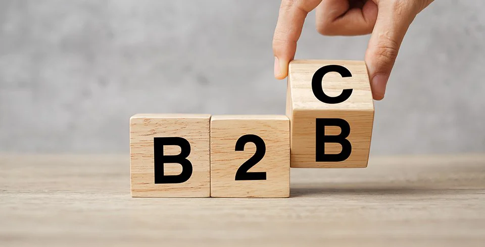 Três blocos de madeira com as letras B2B sendo reorganizados por uma mão que ergue o bloco com a letra C, simbolizando a transição entre os modelos de negócio B2B e B2C.