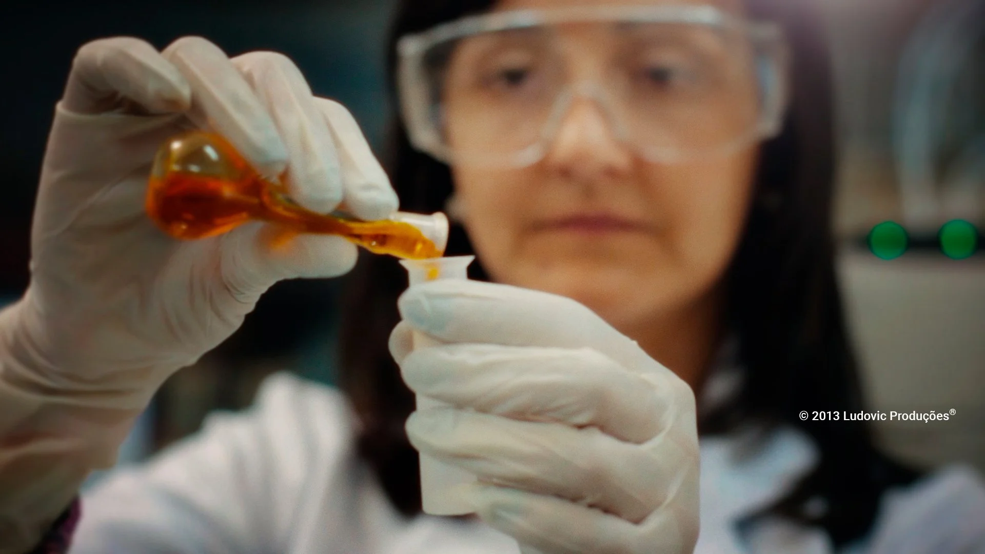 Profissional com luvas brancas manuseia pipeta com líquido laranja em ambiente laboratorial, representando precisão e segurança em vídeo institucional.