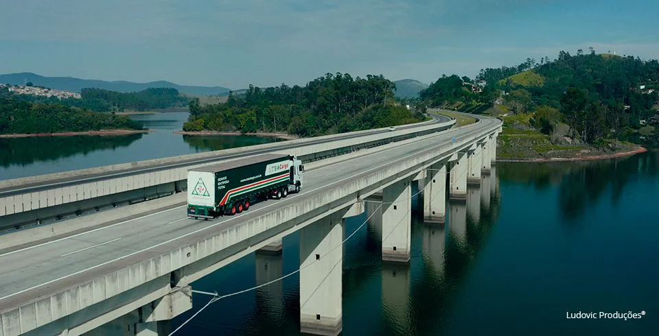 Caminhão da TSA Cargo atravessa ponte sobre rio com colinas verdes ao fundo, simbolizando infraestrutura e presença no vídeo institucional da empresa.