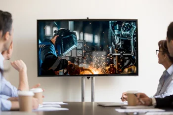 Quatro pessoas assistem à apresentação de vídeo com imagem de soldagem em tela grande durante reunião empresarial, representando uso estratégico de vídeos institucionais.
