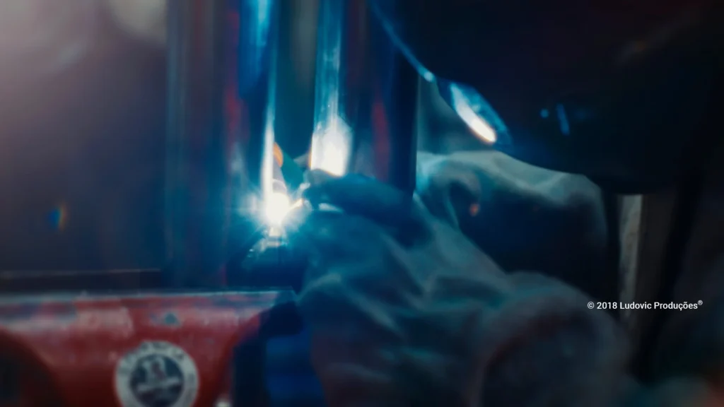 Profissional com capacete de proteção realiza solda em peça metálica, com faíscas visíveis, representando especialização técnica e segurança no ambiente industrial.