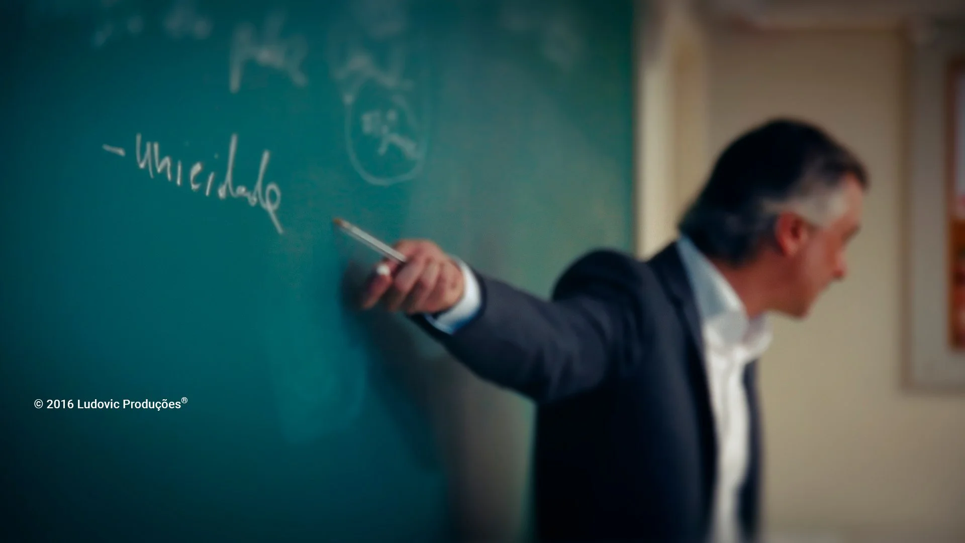 Professor universitário aponta para quadro-negro com a palavra 'unicidade' escrita, simbolizando ensino superior e comunicação institucional em universidades.