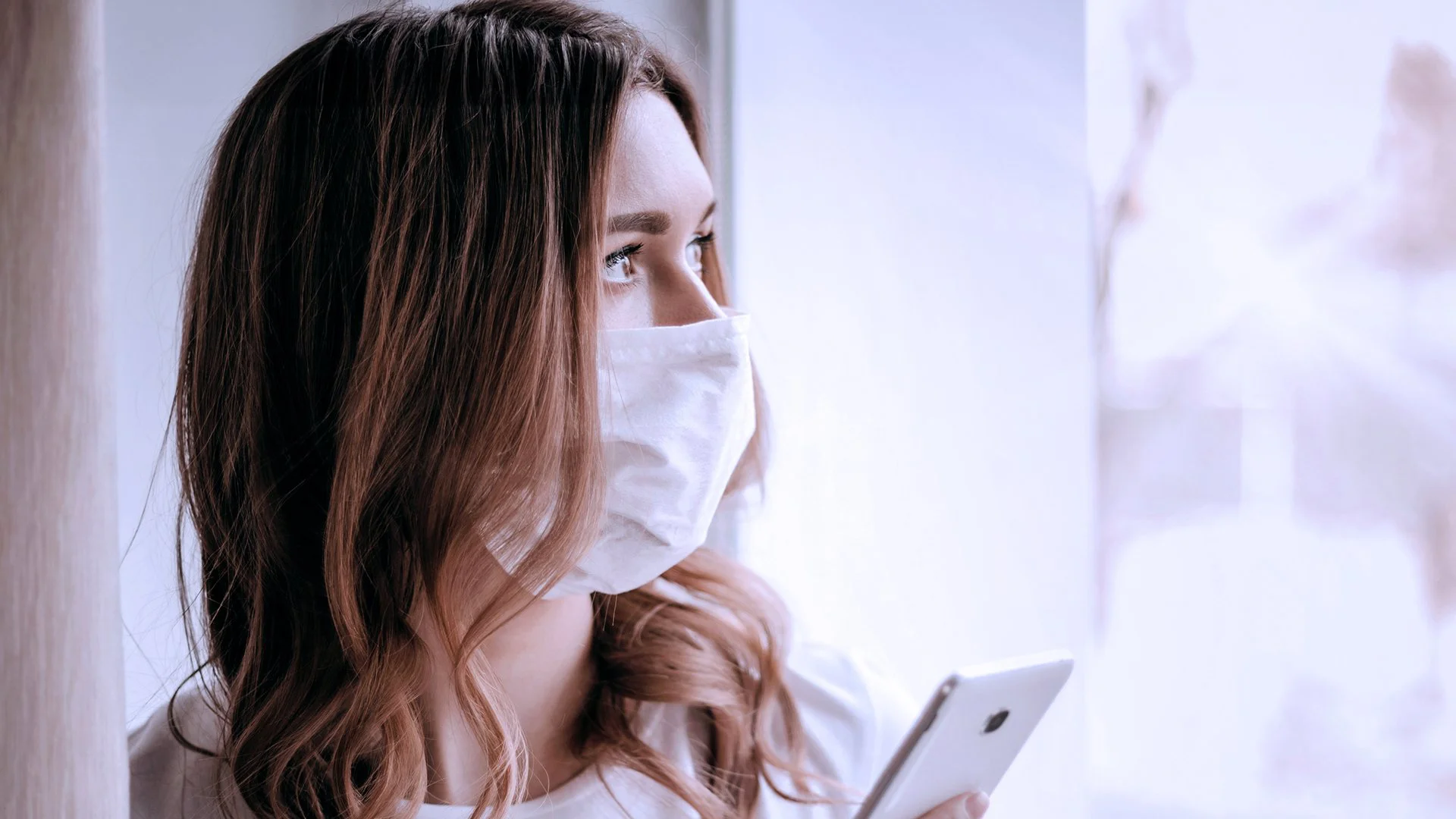 Mulher de máscara usando smartphone perto da janela, com luz suave entrando, simbolizando conexão digital durante o distanciamento social.