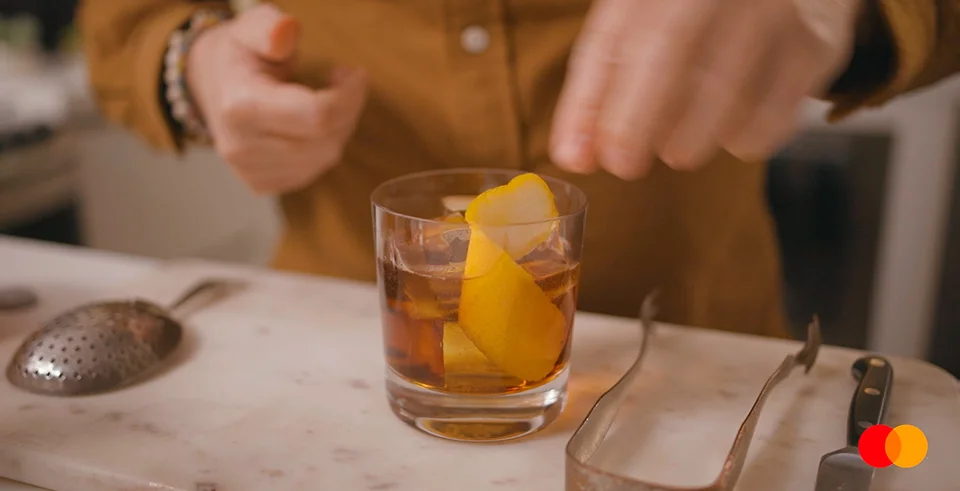 Pessoa coloca guarnição em drink com gelo em copo baixo sobre bancada de mármore, simbolizando ritual e sofisticação na preparação de coquetel.