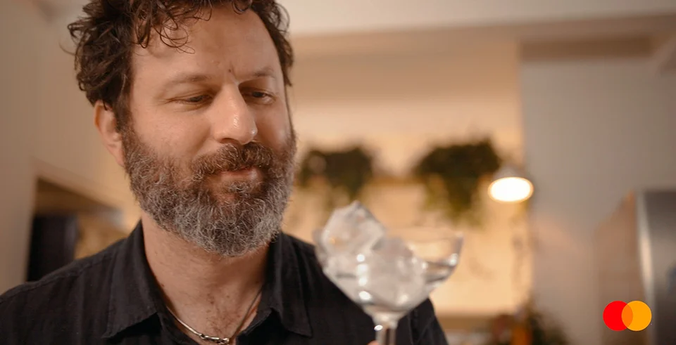 Close em mão segurando copo com gelo em ambiente de cozinha moderna, com plantas ao fundo e logo da Mastercard no canto, representando sofisticação e cotidiano no episódio sobre drinks.