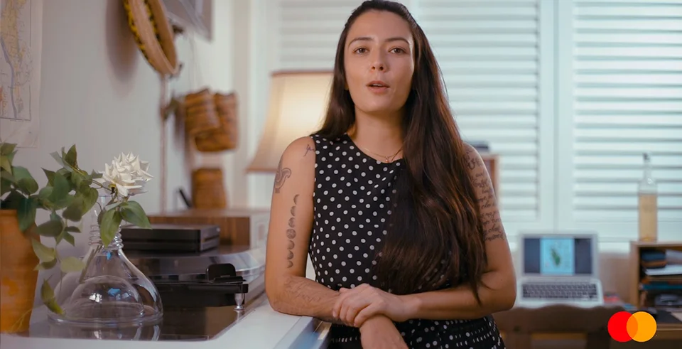 Gabi Monteleone, sommelière, em ambiente acolhedor com objetos afetivos e garrafa de vinho sobre a estante, em websérie para a Mastercard.