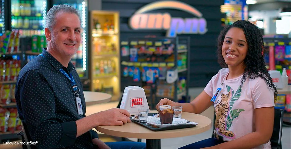 Duas pessoas sentadas em mesa com copos de café em loja da ampm, em ambiente descontraído com prateleiras e logo da marca. Bastidor de produção de vídeo institucional.