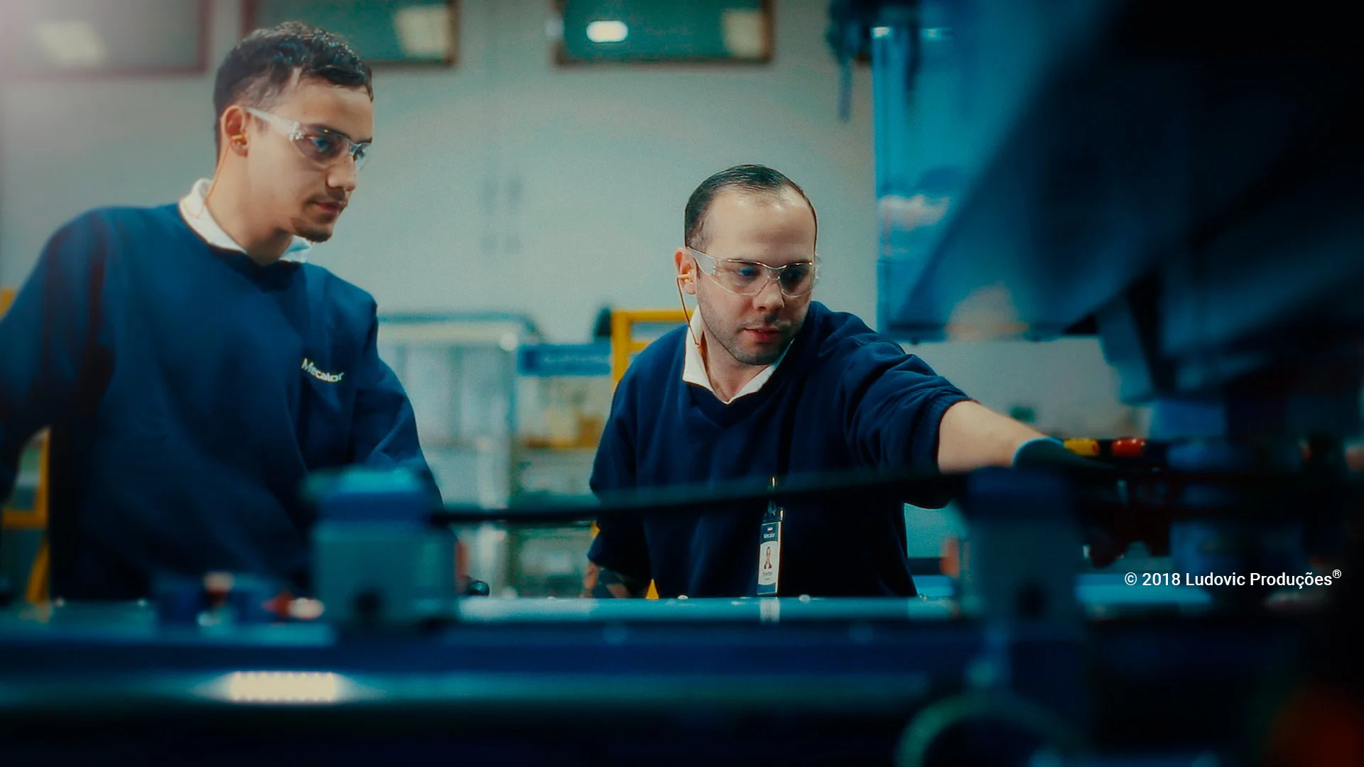 Dois profissionais em uniformes azuis e proteção auricular operando máquina em ambiente industrial, representando excelência e profissionalismo em vídeo institucional.