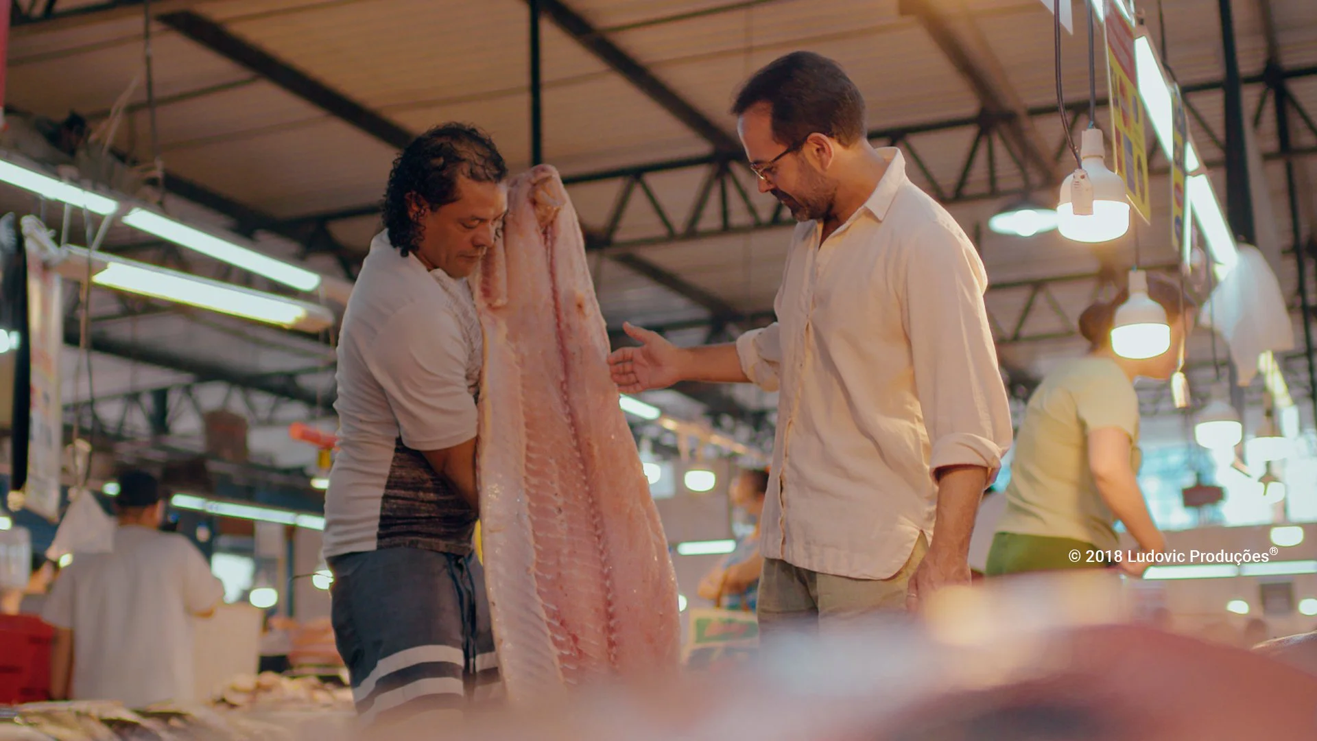 Chef Onildo Rocha interage com vendedor de peixes em mercado amazônico, simbolizando a cultura do pescado e o saber local na gastronomia.
